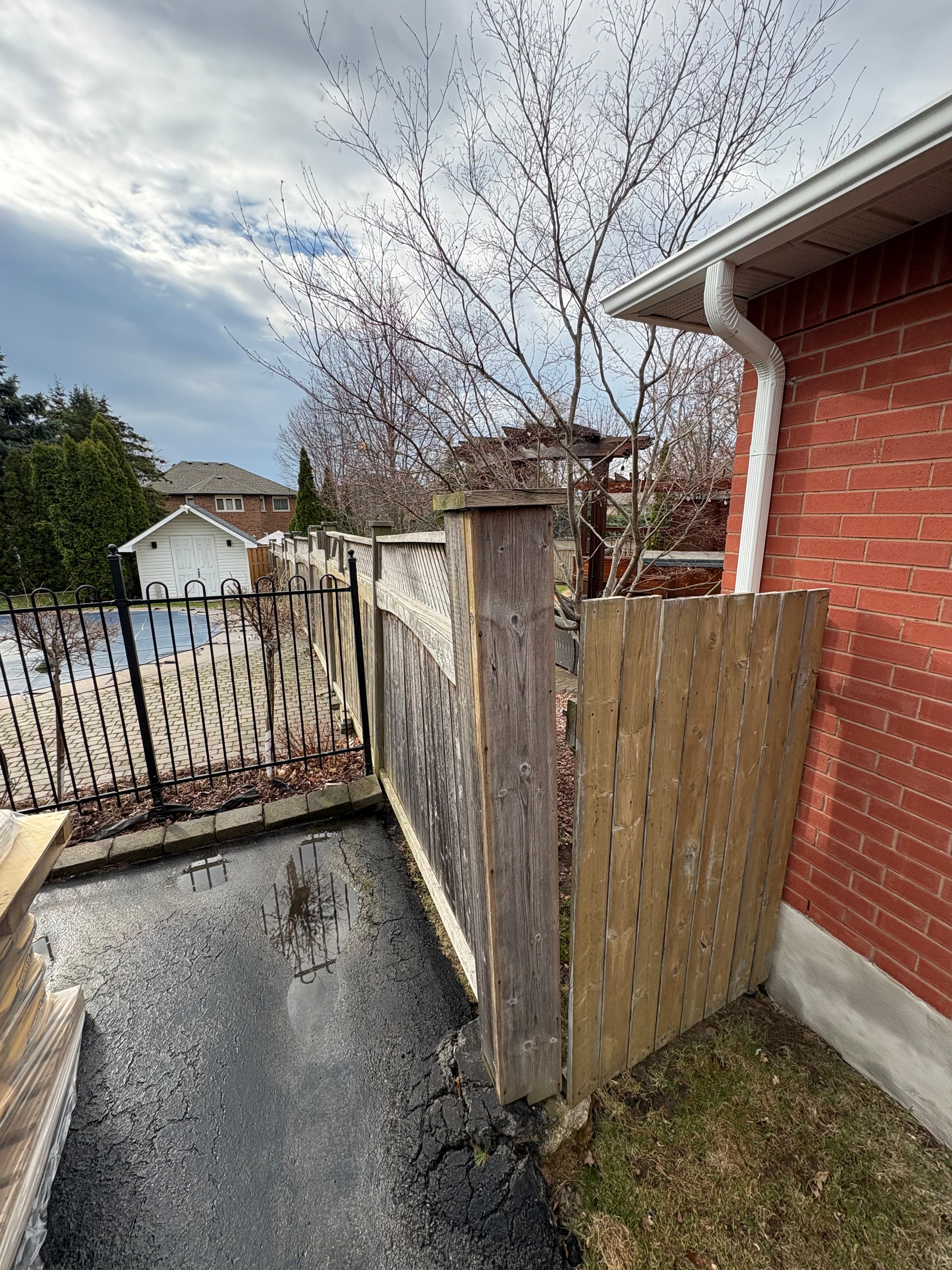 Old weathered grey wood fence and short privacy screen beside a brick house with water pooling on the driveway in Burlington before replacement